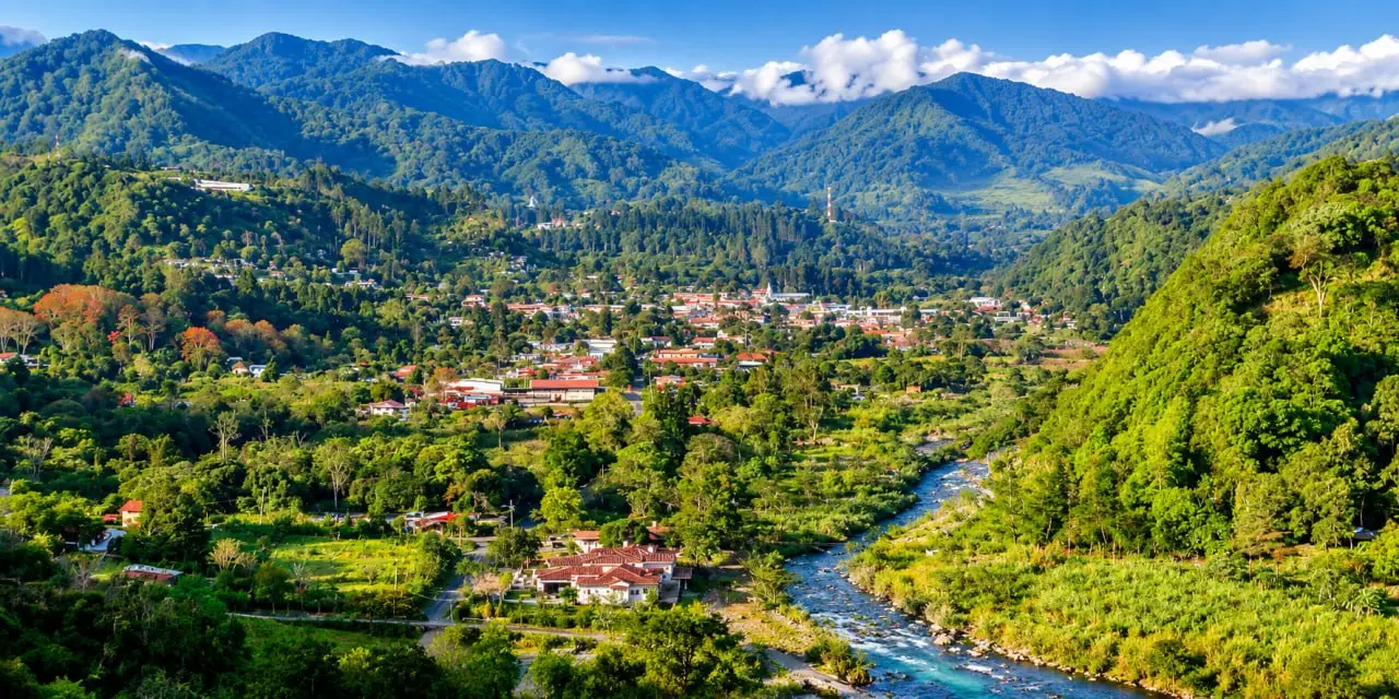 A turquoise river in a wooded gorge, houses on a terrace of the slope, cloud along the ridges