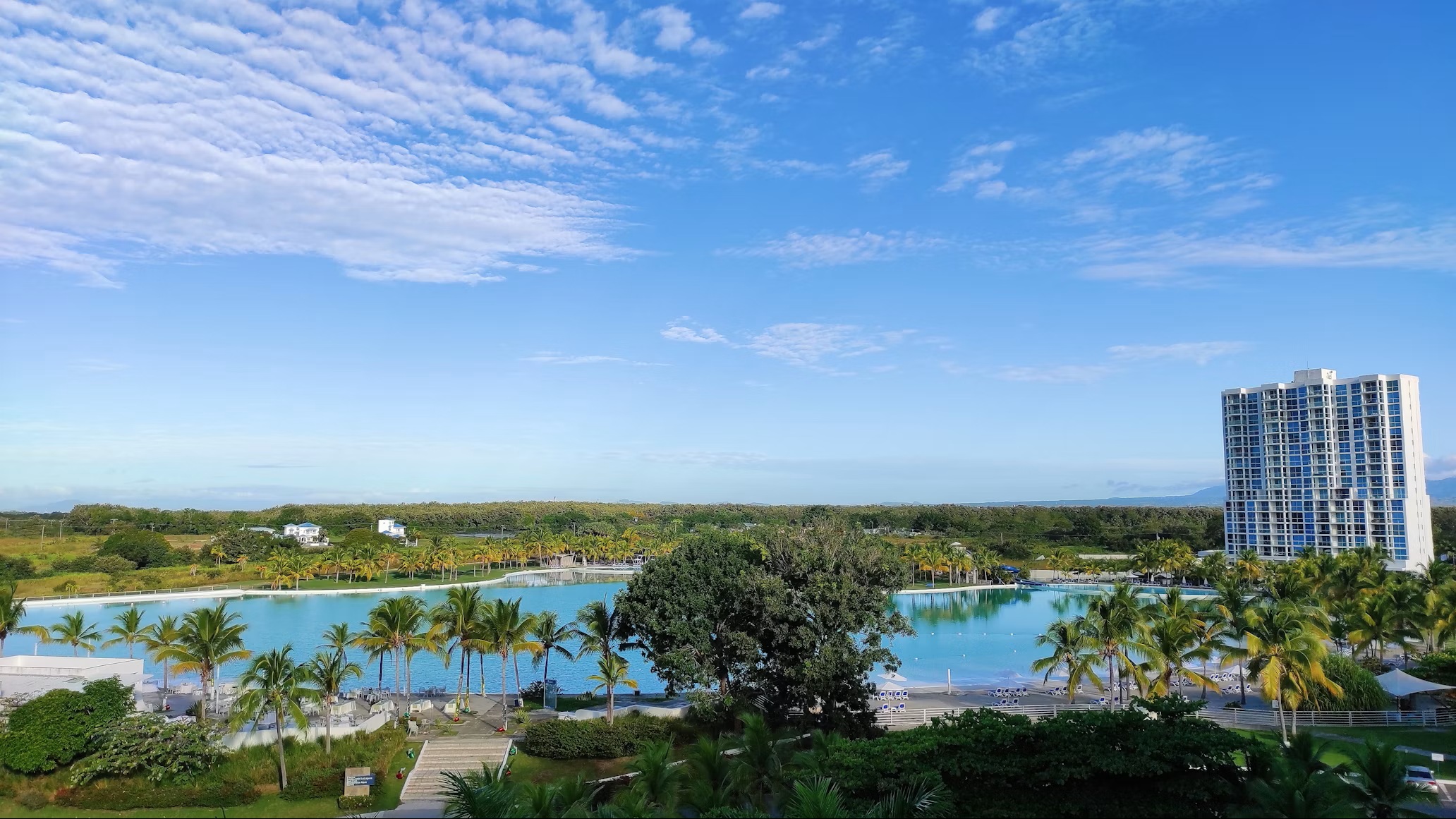 From above: a turquoise lagoon ringed by palms, a tall residential tower to the right, the plain and the hills beyond