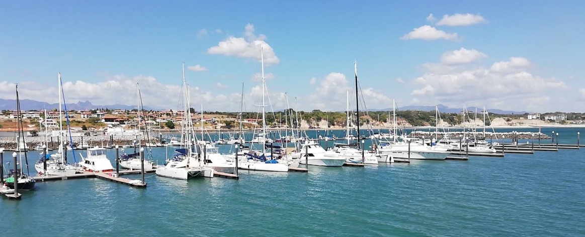 Yachts and motor boats at the pontoons of a marina, a breakwater and a low shore with houses behind
