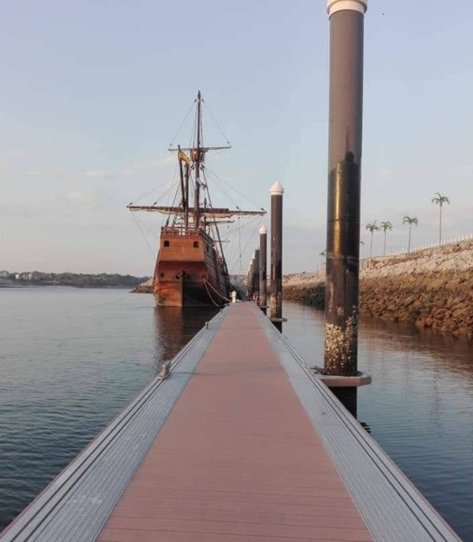 A pontoon walkway leading to a wooden sailing ship with masts, a rock revetment to the right