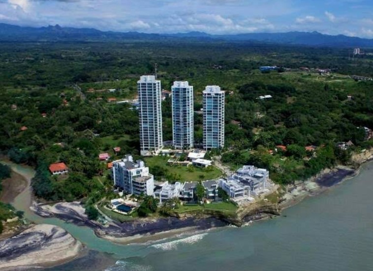 From above: three residential towers and low-rise buildings among greenery on a headland, a river mouth below