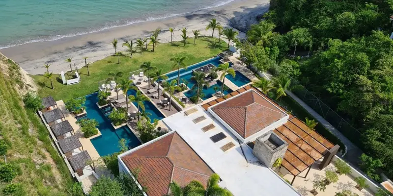 Vista desde lo alto de un club de playa: una piscina larga con tumbonas, tejados de teja, detrás arena gris